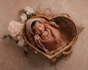 Smiling newborn baby wrapped in pink, sleeping in a heart-shaped wicker basket with flowers.