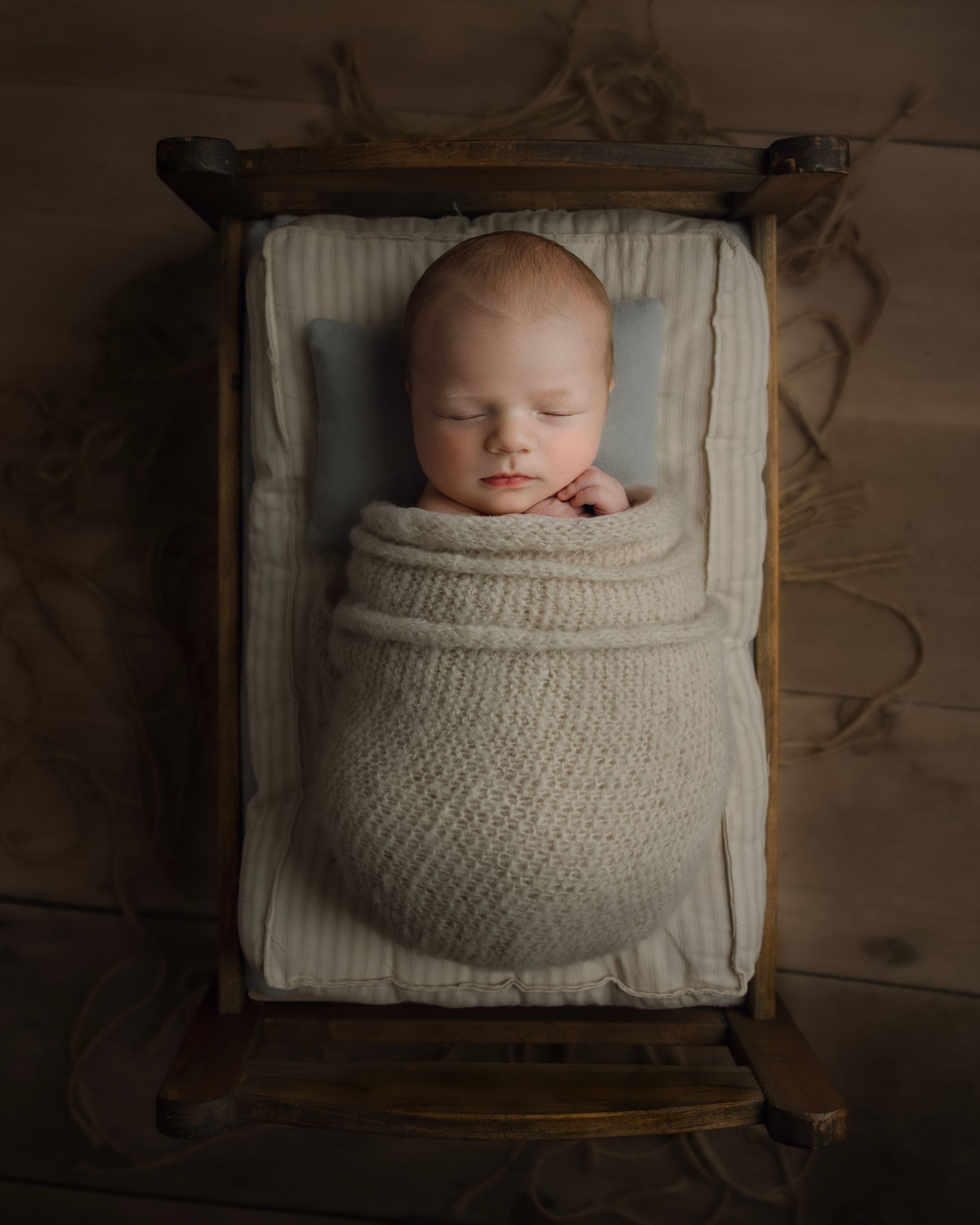 Sleeping newborn baby swaddled in cream knit wrap, resting peacefully in a rustic wooden mini bed.