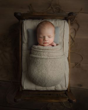 Sleeping newborn baby swaddled in cream knit wrap, resting peacefully in a rustic wooden mini bed.