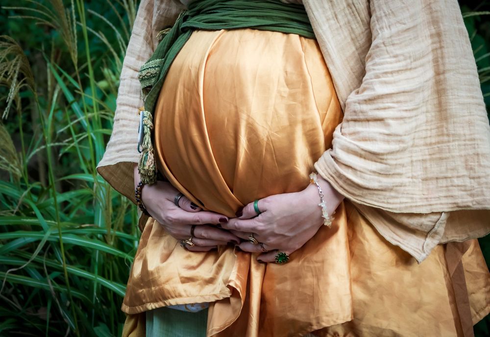 A pregnant belly in a golden dress is tenderly held against a green natural backdrop.