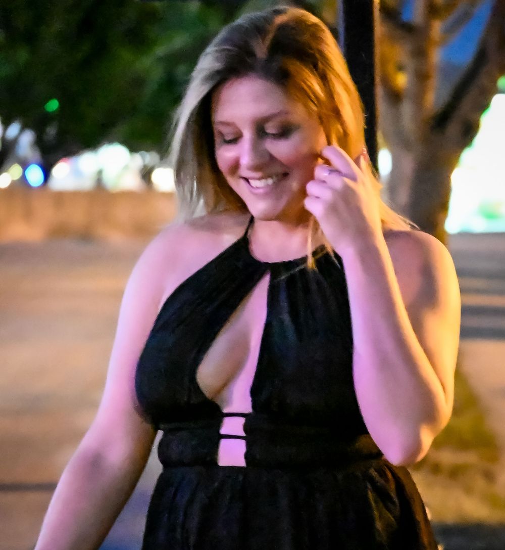 Woman in elegant black dress posing under evening lights with a warm glow.