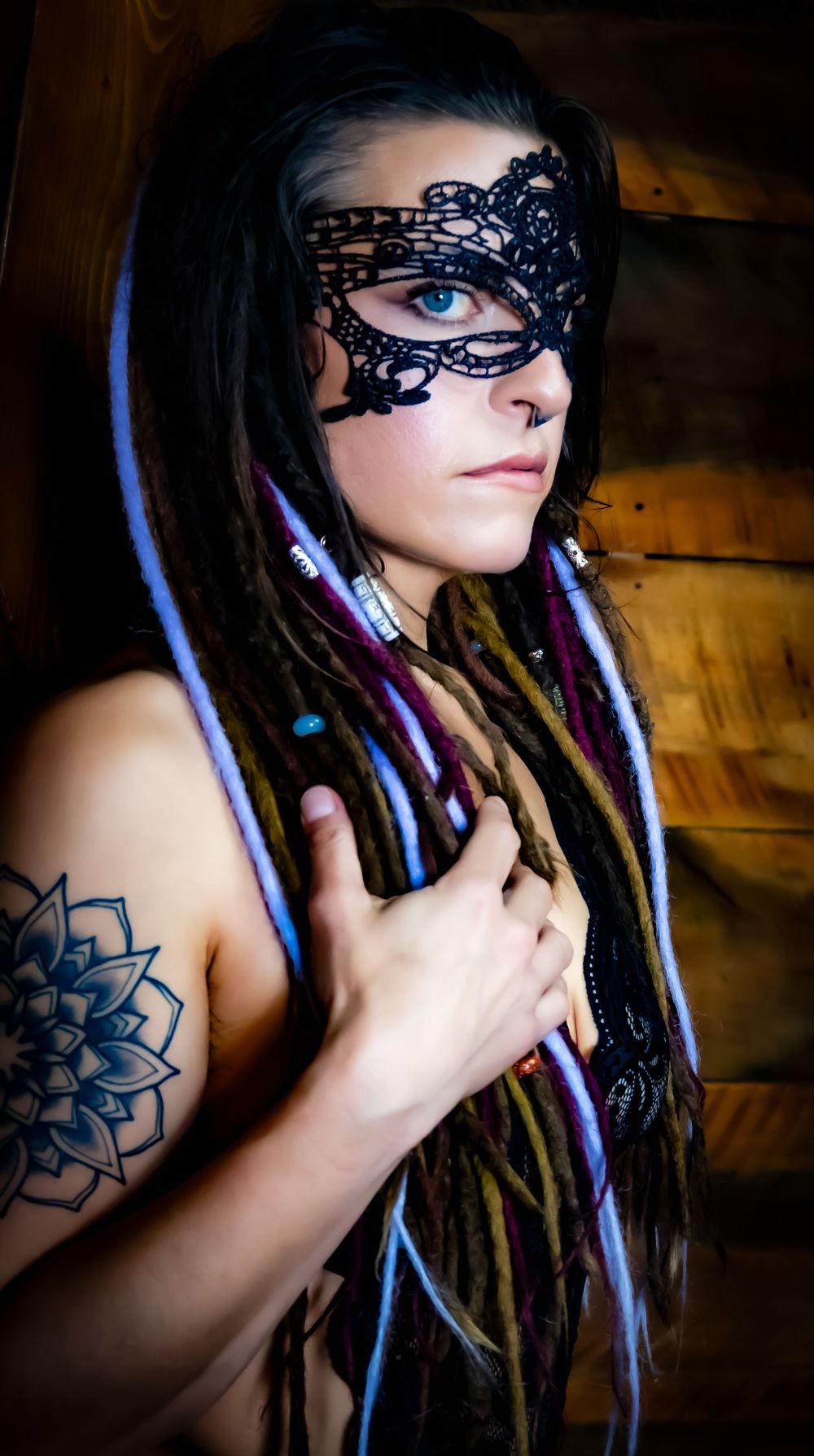 A person wearing a black lace mask with dreadlocks and a visible mandala tattoo poses against a wooden wall.