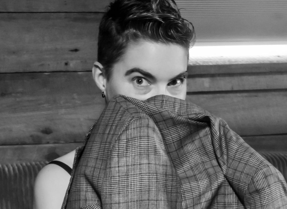 A black and white portrait shows someone playfully hiding behind plaid fabric while gazing at the camera.