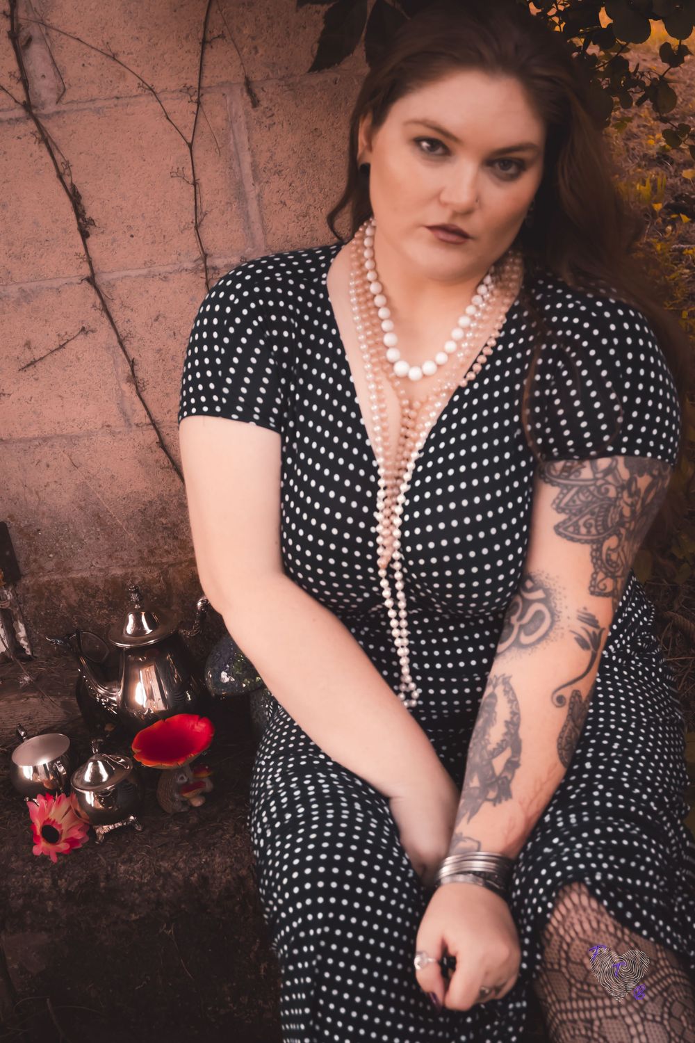 A person models in a vintage-style polka dot dress with pearls and tattoos against a rustic backdrop with wine.