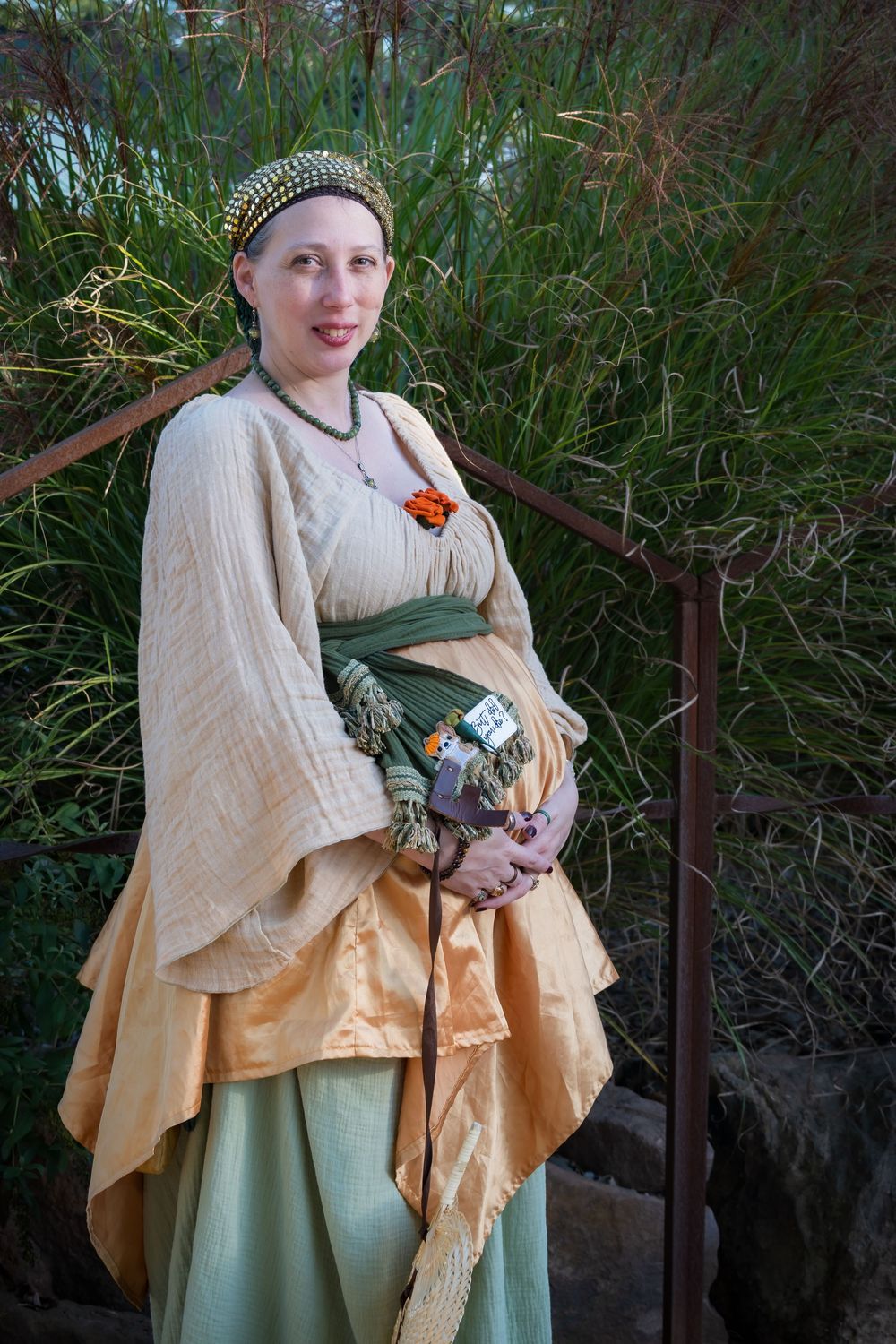 Medieval-style costume features cream shawl, sage green dress and peach layers with decorative brooch in green garden setting.