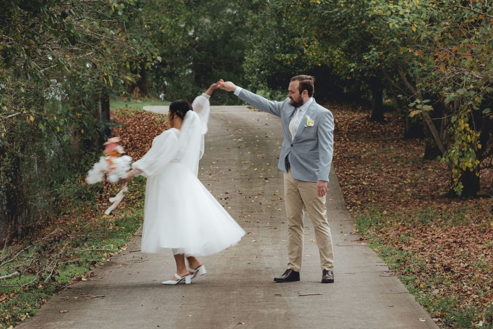 Joyful wedding couple dancing on tree-lined path in casual bridal attire.