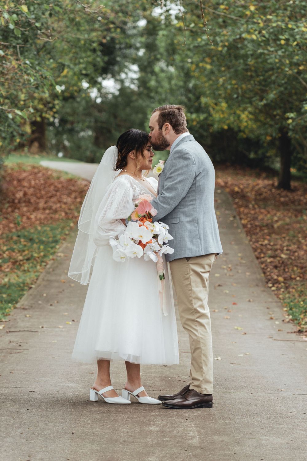 Newlyweds share romantic moment on autumn path with bride holding floral bouquet.