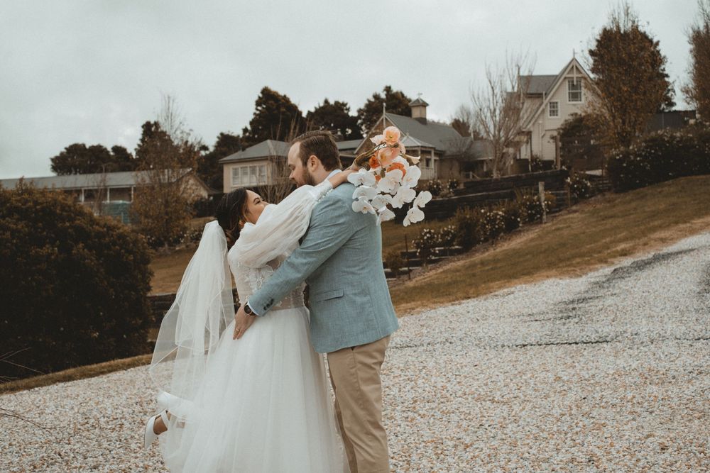 Wedding couple embracing on a rocky beach with coastal homes in the background.
