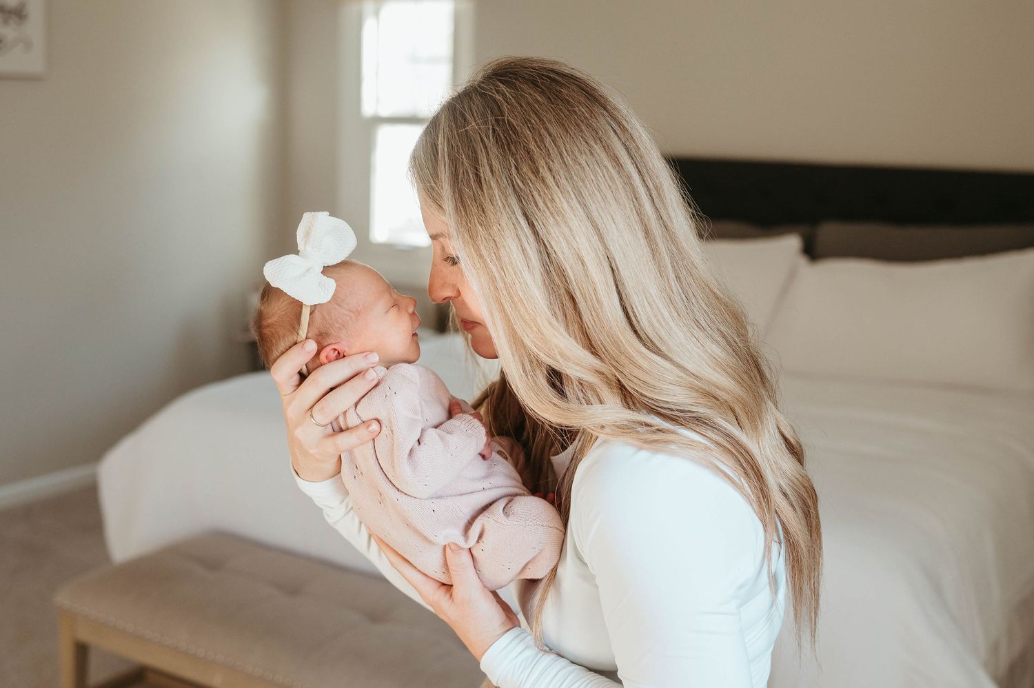 Blonde mother in white top gently kisses her newborn baby dressed in pink in a softly lit bedroom.