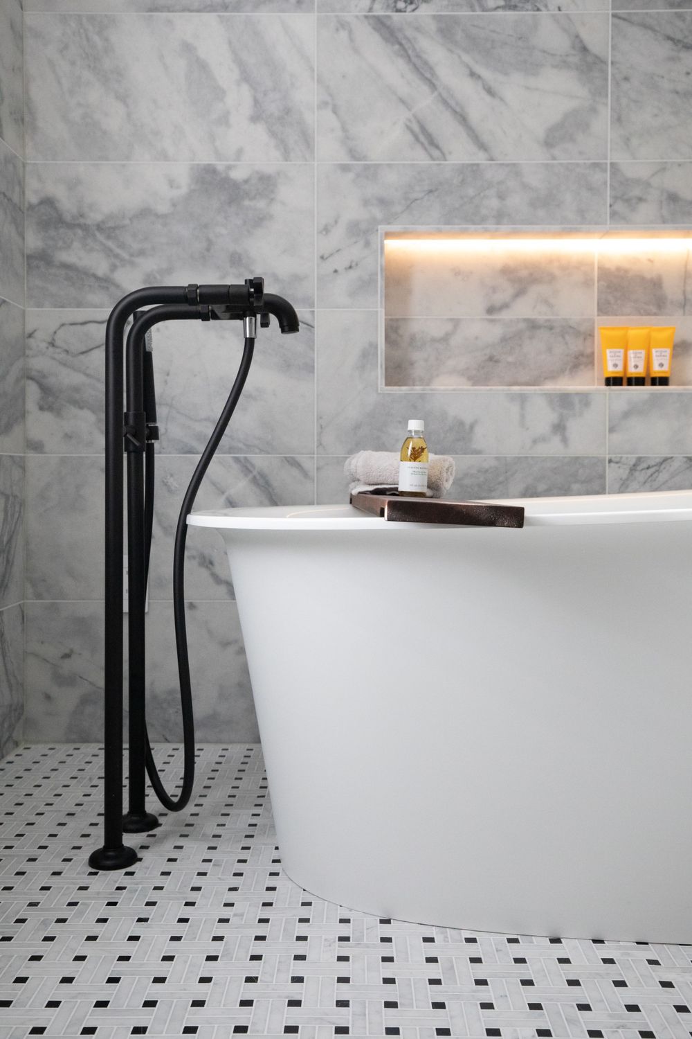 Modern freestanding bathtub with matte black faucet against marble tile walls.