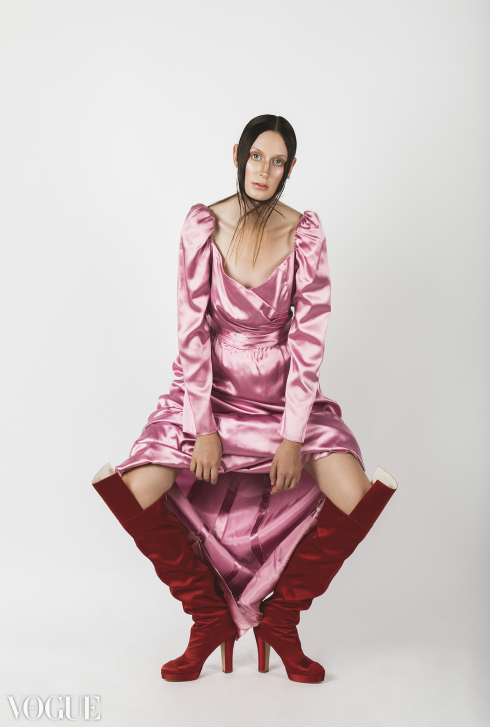 Fashion model in pink satin dress and red boots poses against white background for Vogue.