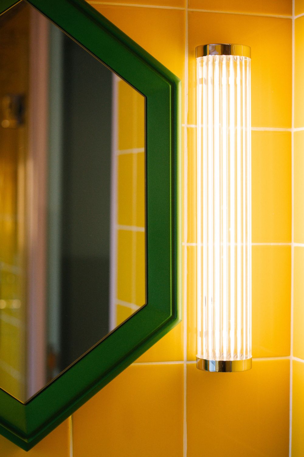 Art deco style wall sconce with ribbed glass shade mounted on yellow tiled wall with green mirror frame.