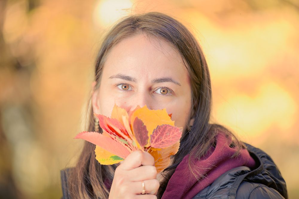 Person holding large orange maple leaf in front of face during autumn season.