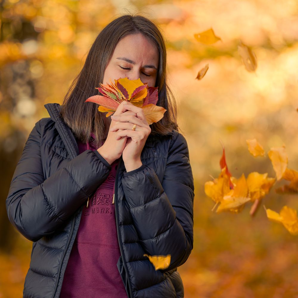 Woman in black puffer jacket smells autumn maple leaf against golden bokeh background in fall.