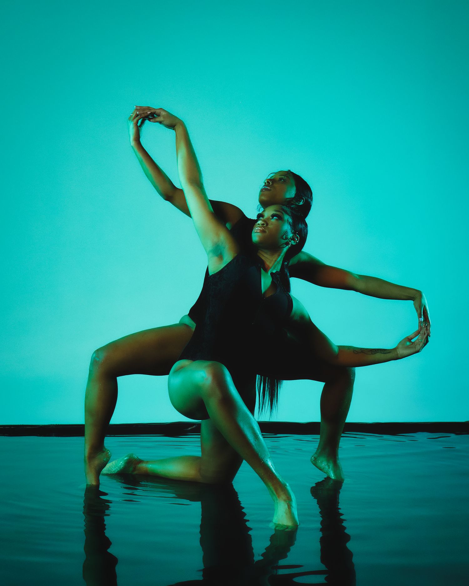 Two dancers perform an artistic duet against a turquoise backdrop on a reflective floor.