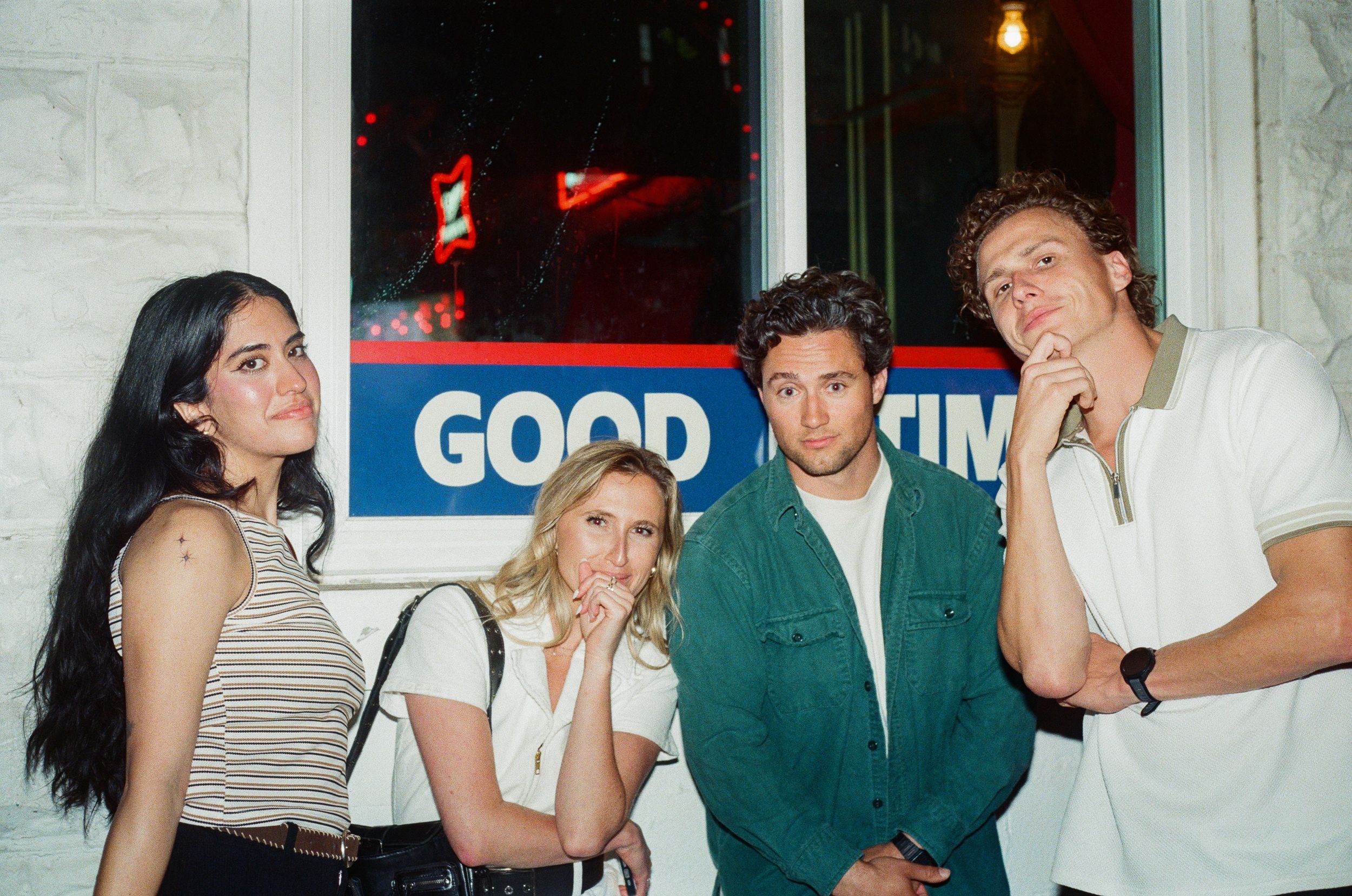Four young people posing casually at night in front of illuminated Goodyear sign with vintage film aesthetic.