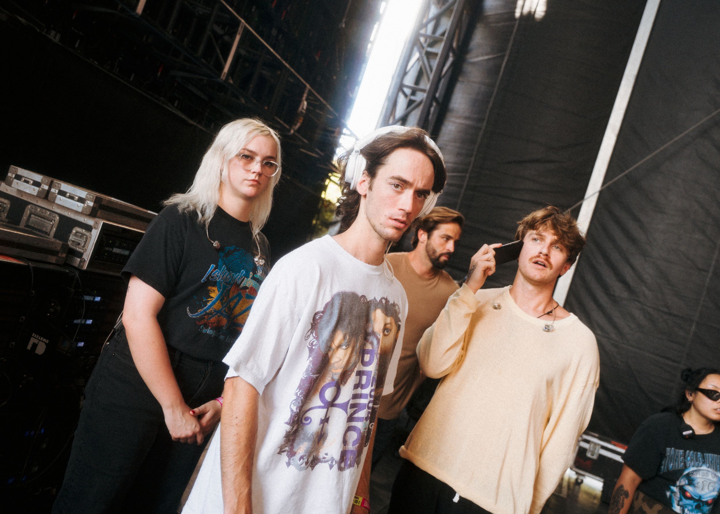Four young band members pose casually backstage with amplifiers and stage equipment visible in the background.