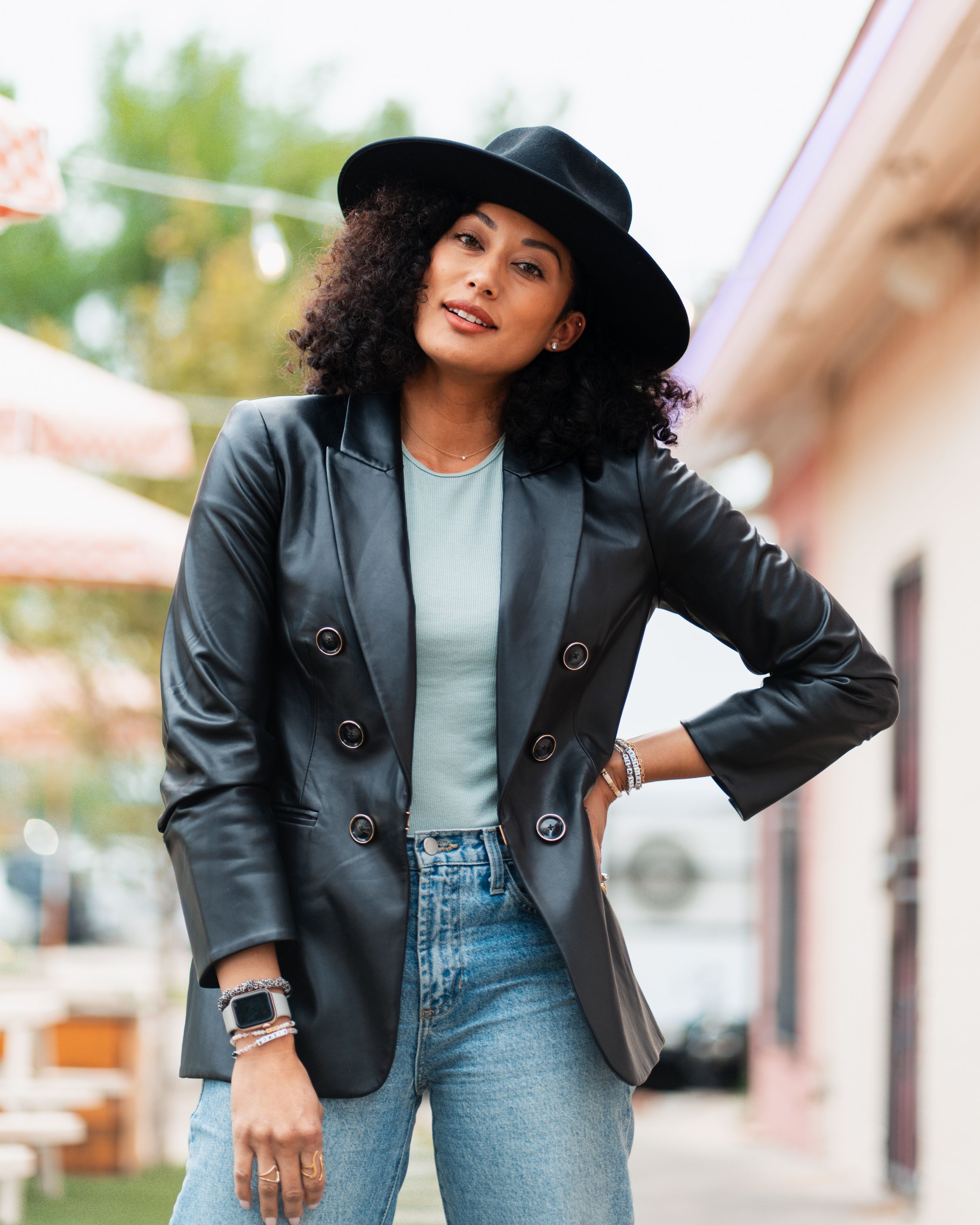 Stylish outfit featuring a black blazer, light gray top, wide-leg jeans and black hat on a sunny street.