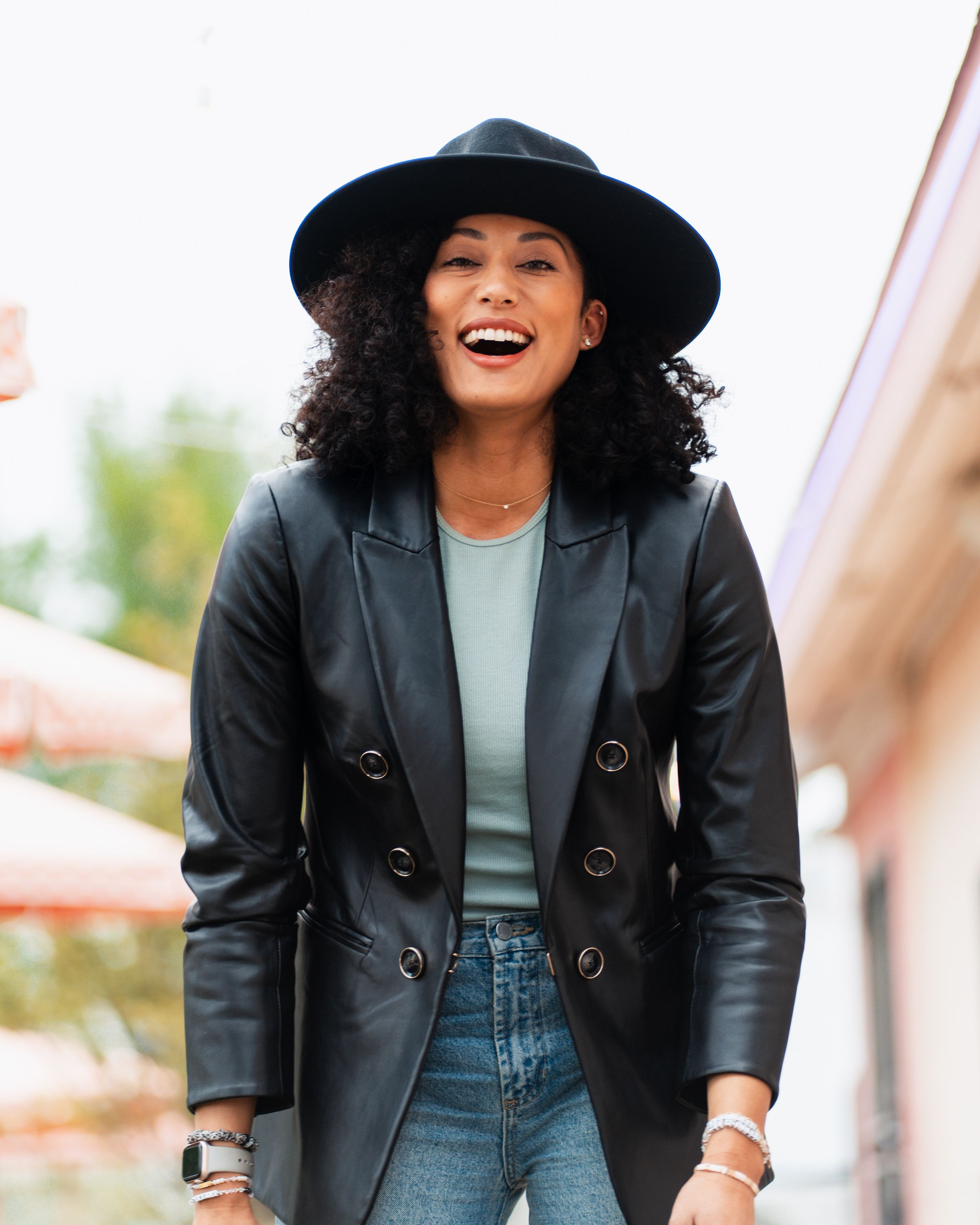 Stylish woman wearing black leather blazer, denim jeans and wide brim hat laughs joyfully outdoors.