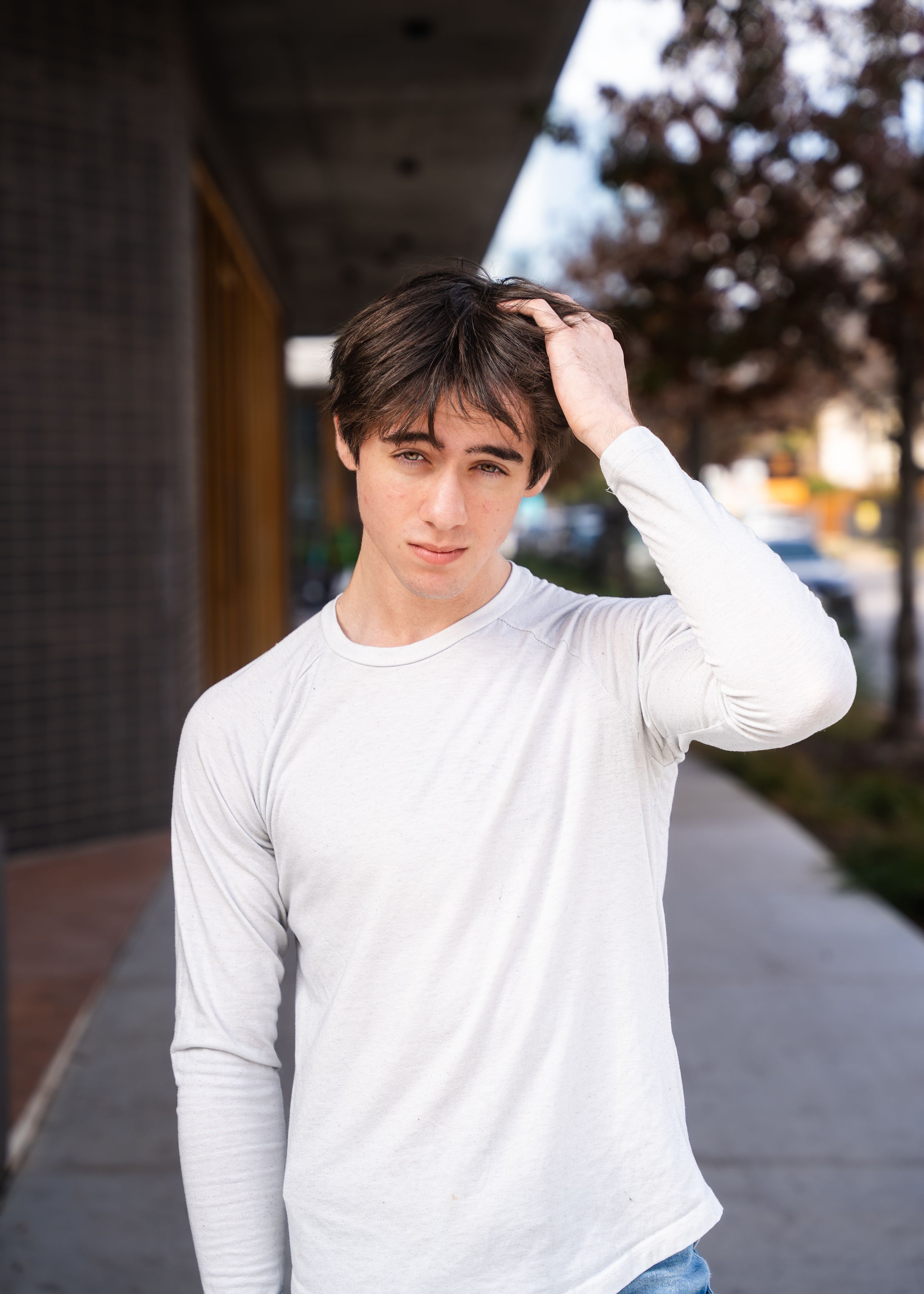 Person wearing a white long sleeve shirt stands on a sidewalk running their hand through their hair at dusk.