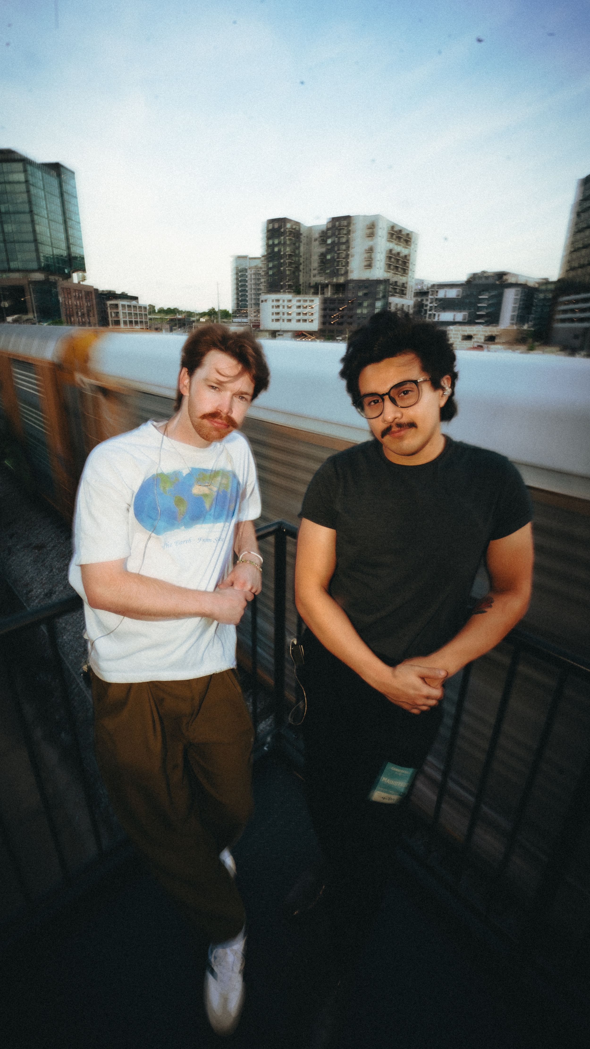 Two friends pose casually together on an urban rooftop deck with city buildings visible in the background.