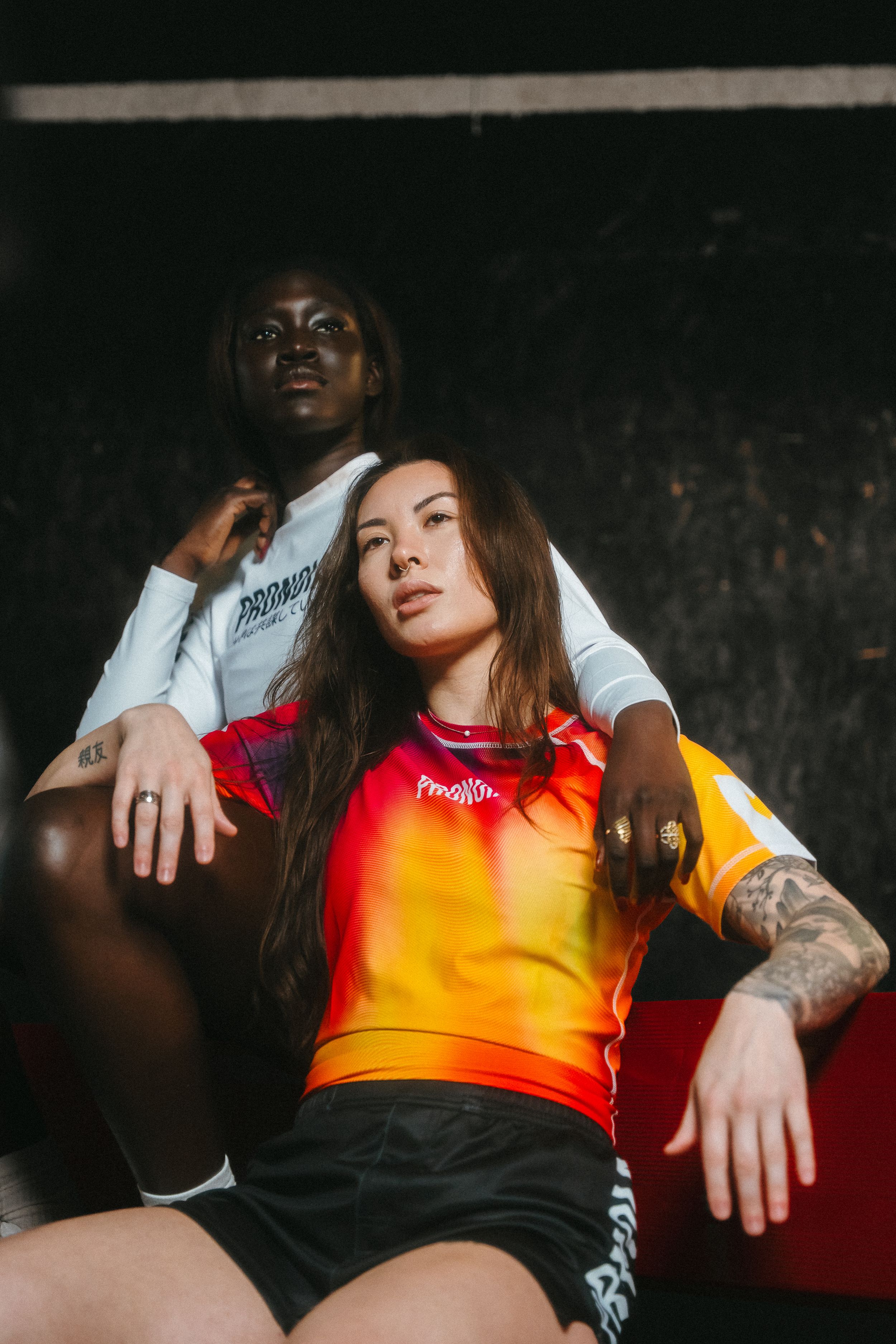 Two athletes in contrasting white and orange-red sportswear pose together against dark backdrop in moody portrait.