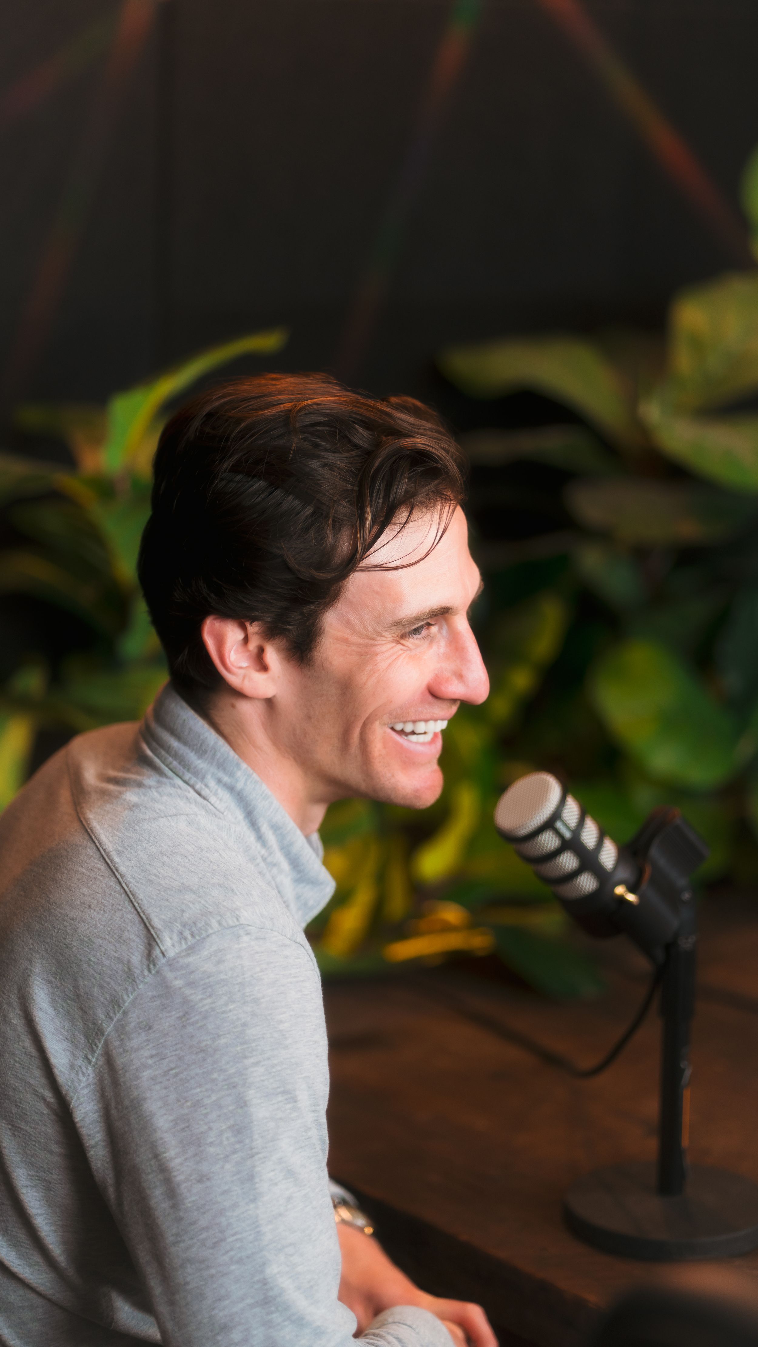 A speaker wearing a gray sweater laughs while recording a podcast episode at a microphone setup.