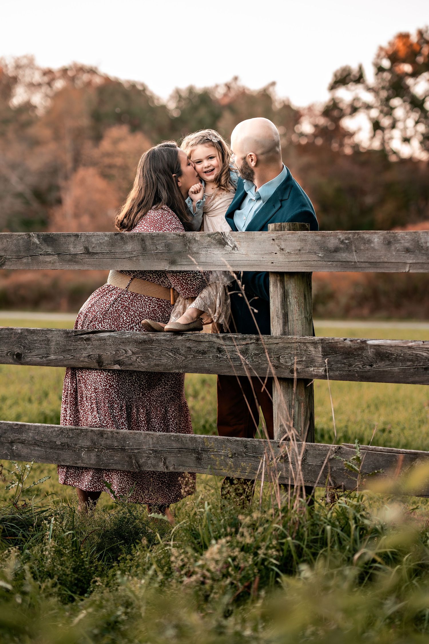 A family shares a tender moment together by a rustic wooden fence during golden hour in an autumn field.