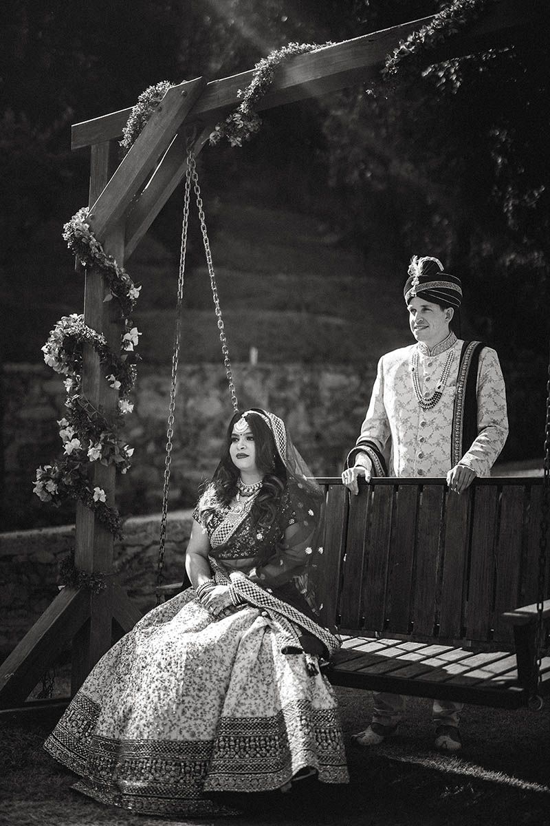 Black and white photography of a couple in traditional Indian wedding attire posing on decorated wooden swing.