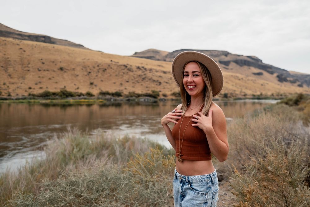 Person in brown top and denim by river wearing wide-brim hat in desert landscape.