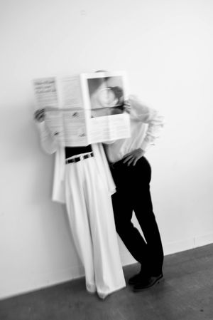 Black and white photo of two people embracing while holding a newspaper covering their faces, wearing contrast outfits.