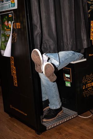 Person wearing jeans and Converse sneakers sitting inside a black photo booth with curtains.