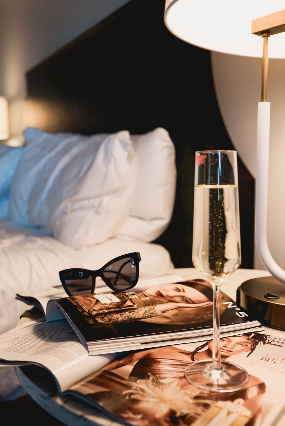 Elegant still life with champagne glass, sunglasses, and fashion magazines on hotel bedding.