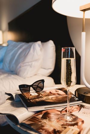 Elegant still life with champagne glass, sunglasses, and fashion magazines on hotel bedding.