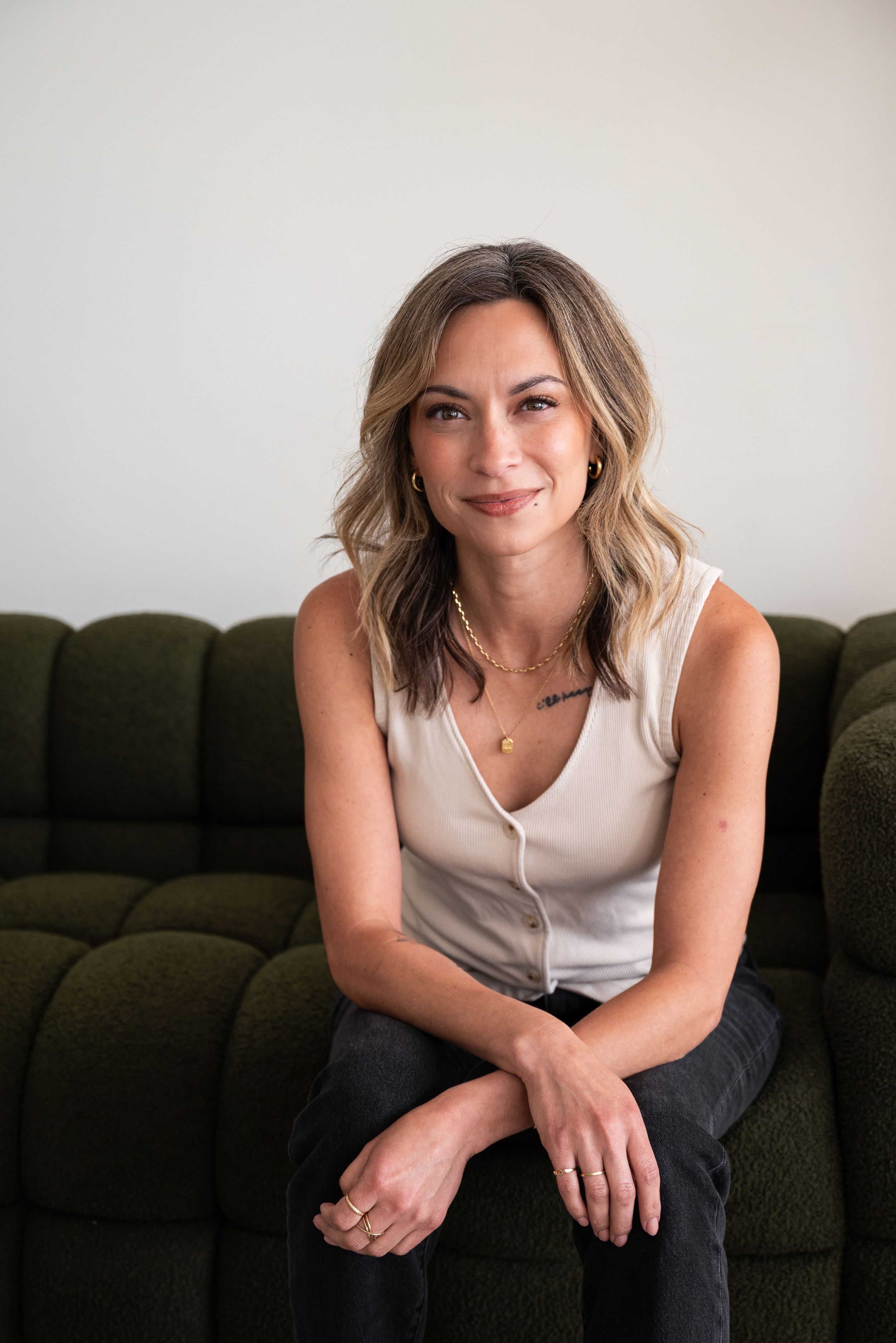 Woman with wavy blonde hair smiling, seated on a dark green textured sofa, wearing a white sleeveless top.