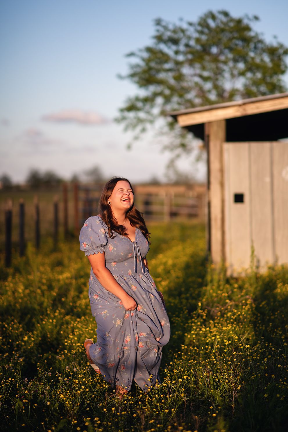 Joyful young woman in blue floral maxi dress laughs in wildflower field at golden sunset.
