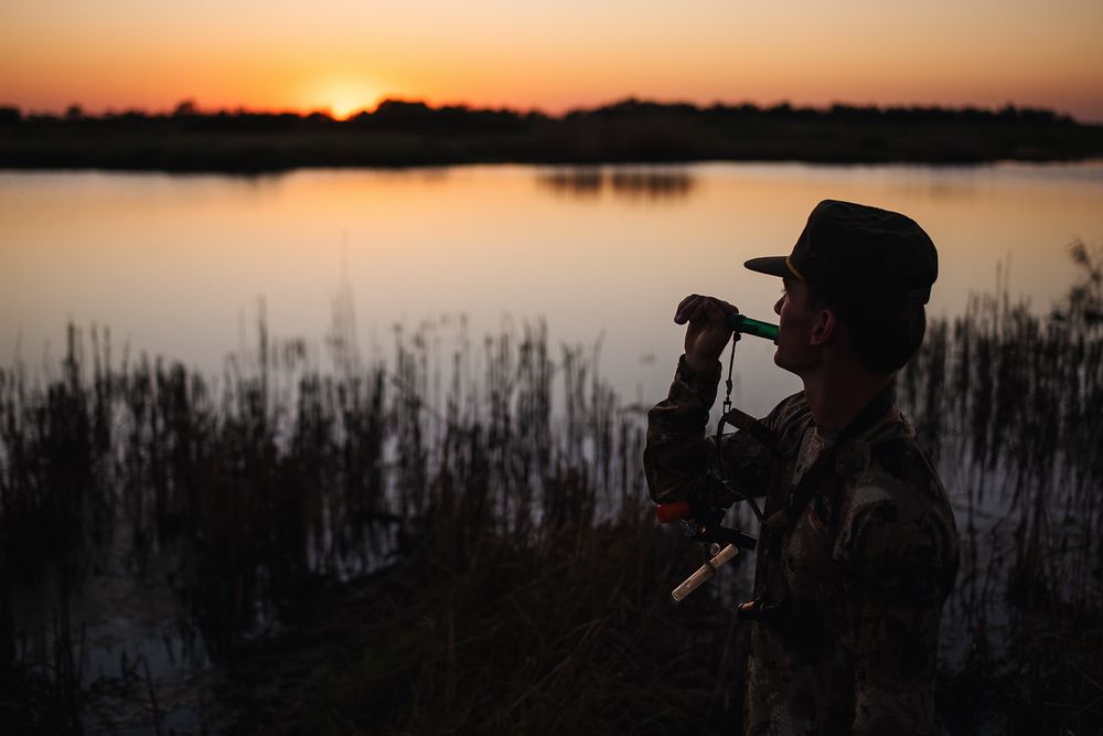 Silhouette of person with hat and camera gear by a serene lake at sunset with warm orange sky.