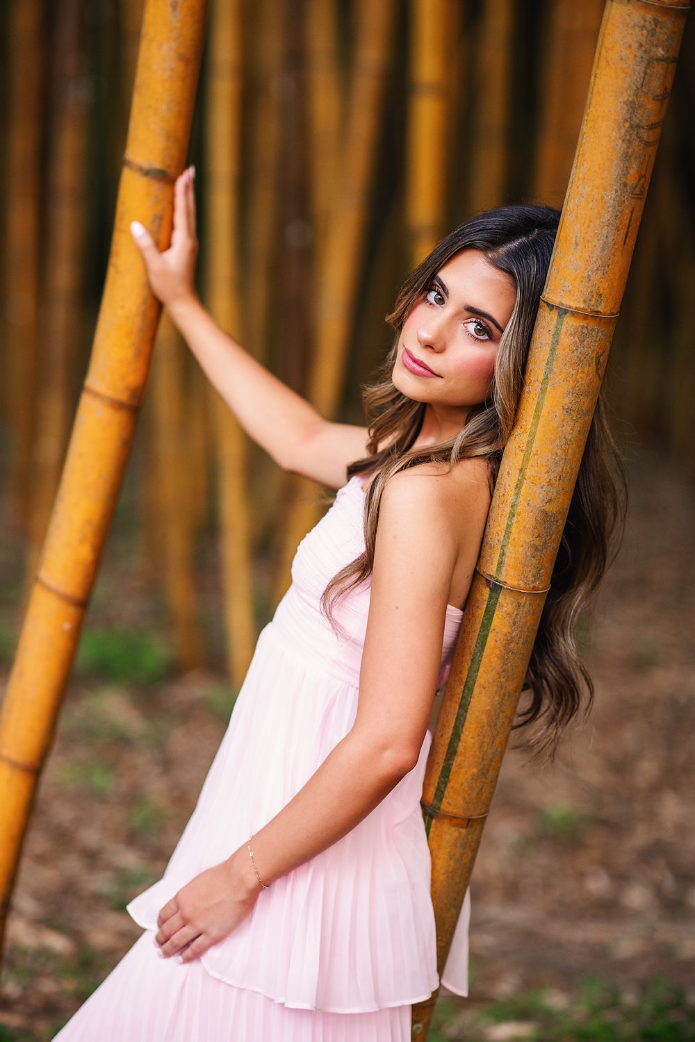 Brunette woman in pink dress posing gracefully between bamboo stalks in forest.