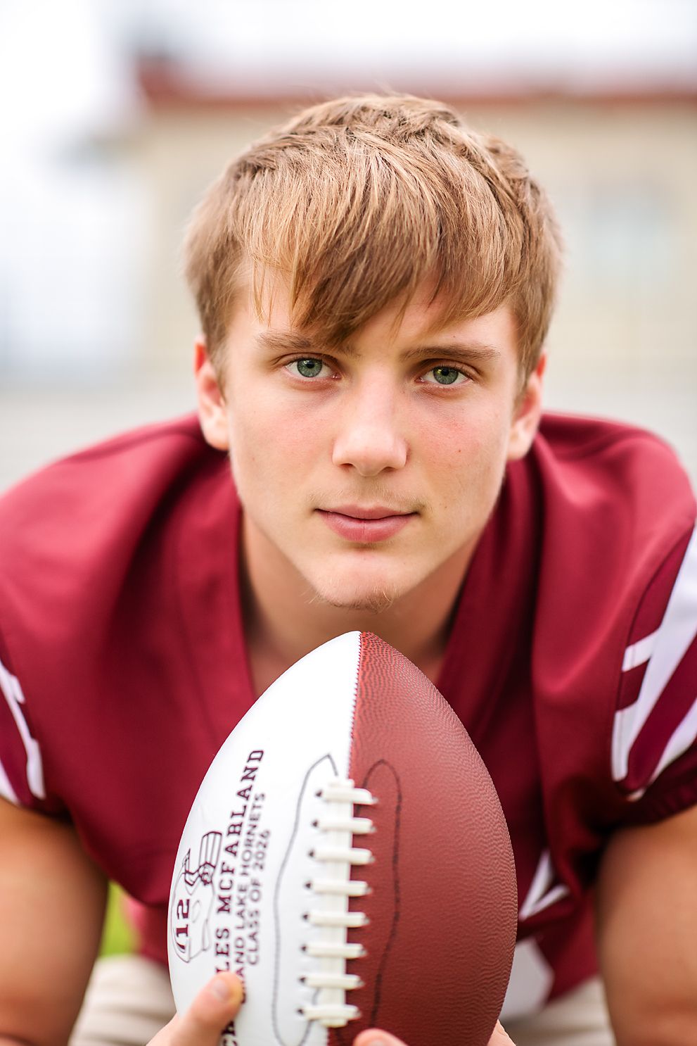 Young male football player in maroon jersey holding football toward camera outdoors.