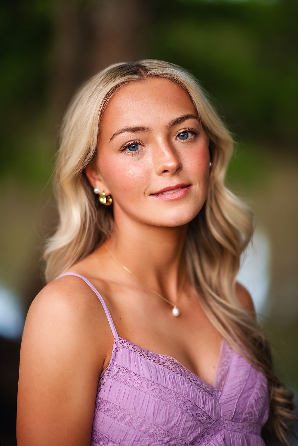Blonde young woman in lavender dress with gold earrings poses for portrait in soft natural light.