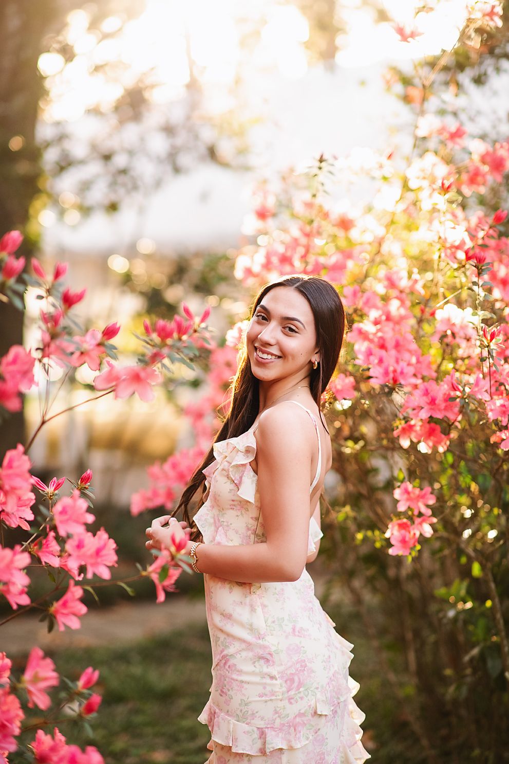 Smiling young woman in floral dress standing among vibrant pink blooming flowers at sunset.