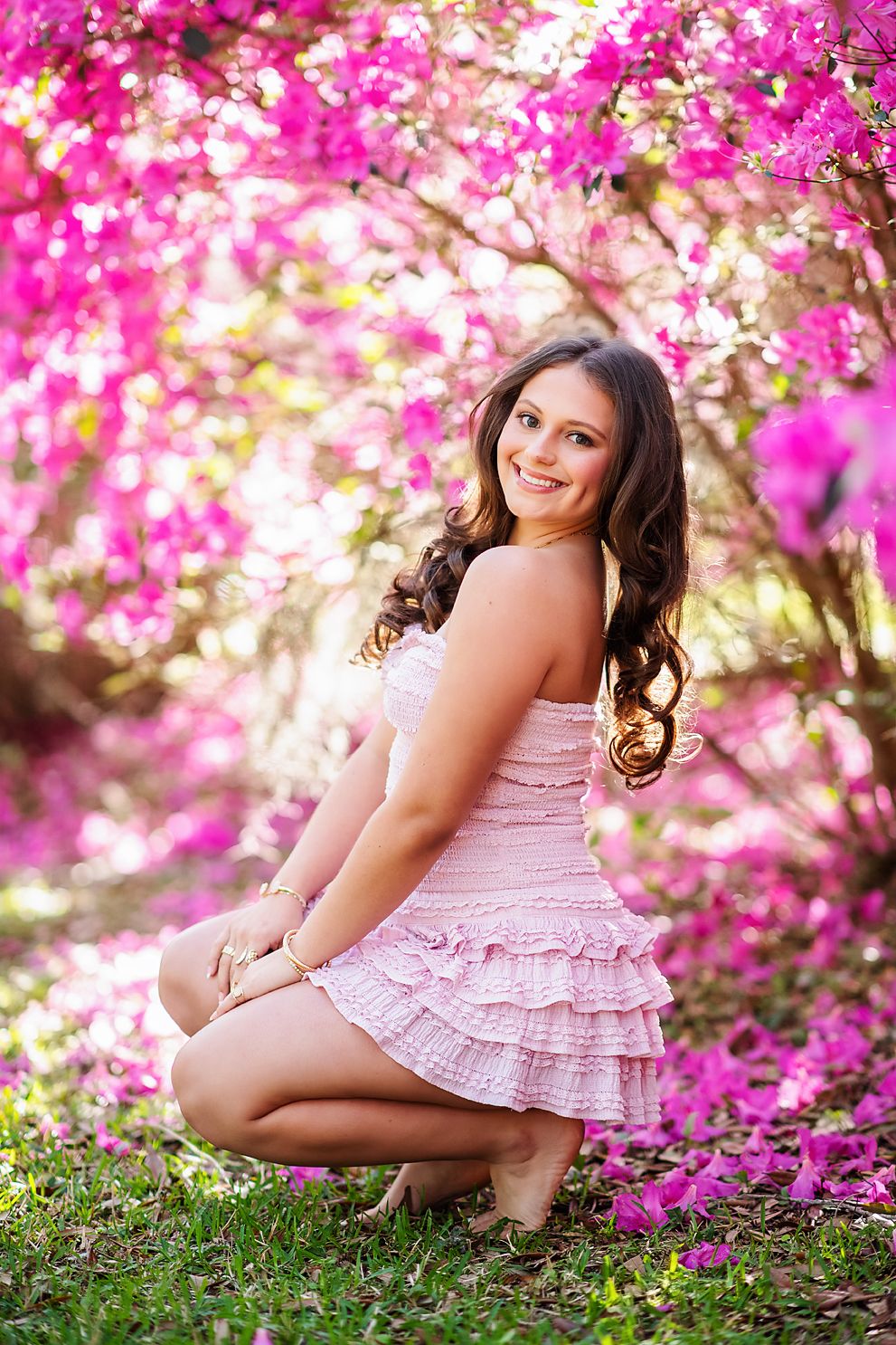 Smiling young woman in pink ruffled dress crouching among vivid pink flowering trees.