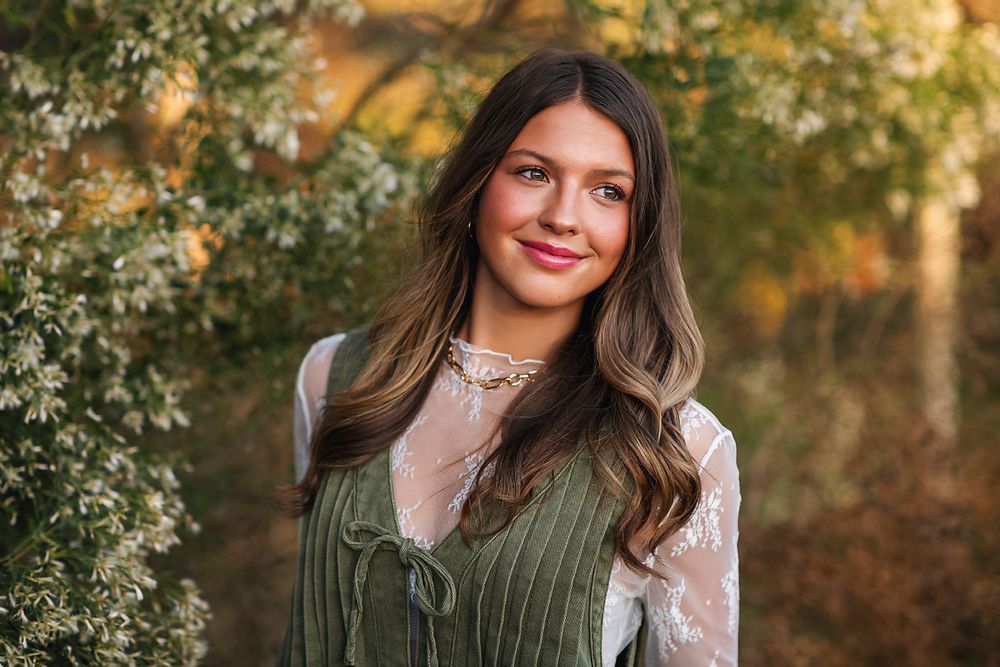 Teenage girl with long brunette hair smiling outdoors near white flowering bushes in warm autumn light.
