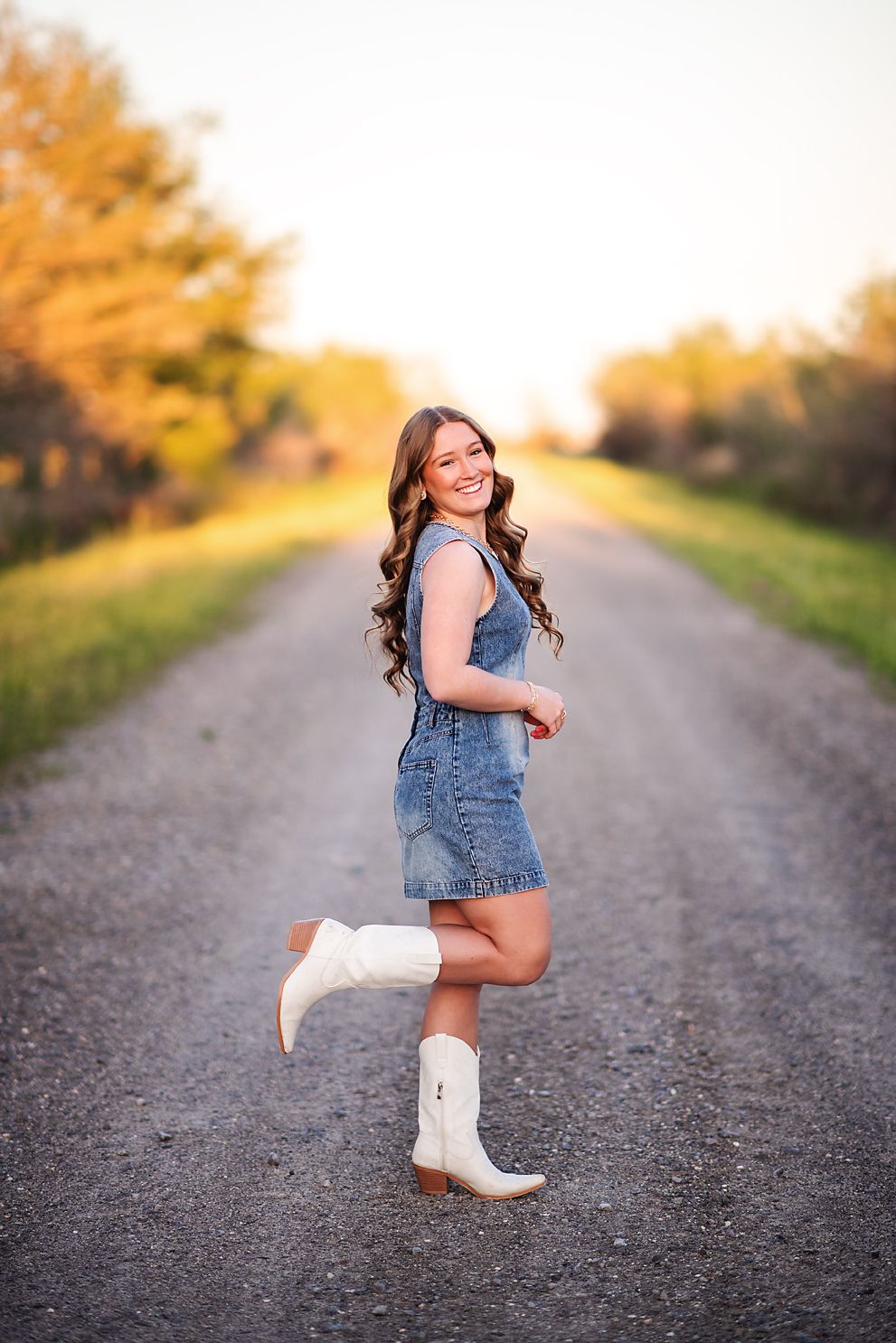 Smiling girl in denim overalls and white cowboy boots posing on rural dirt road at sunset.