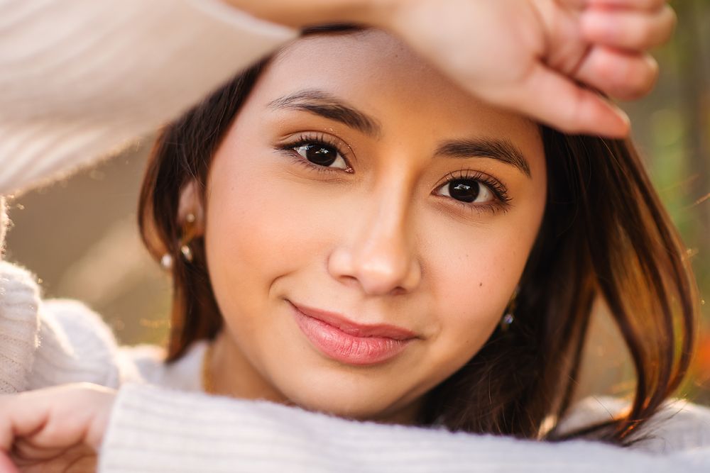 Young Hispanic woman smiling outdoors in warm sunlight, hand raised to forehead.