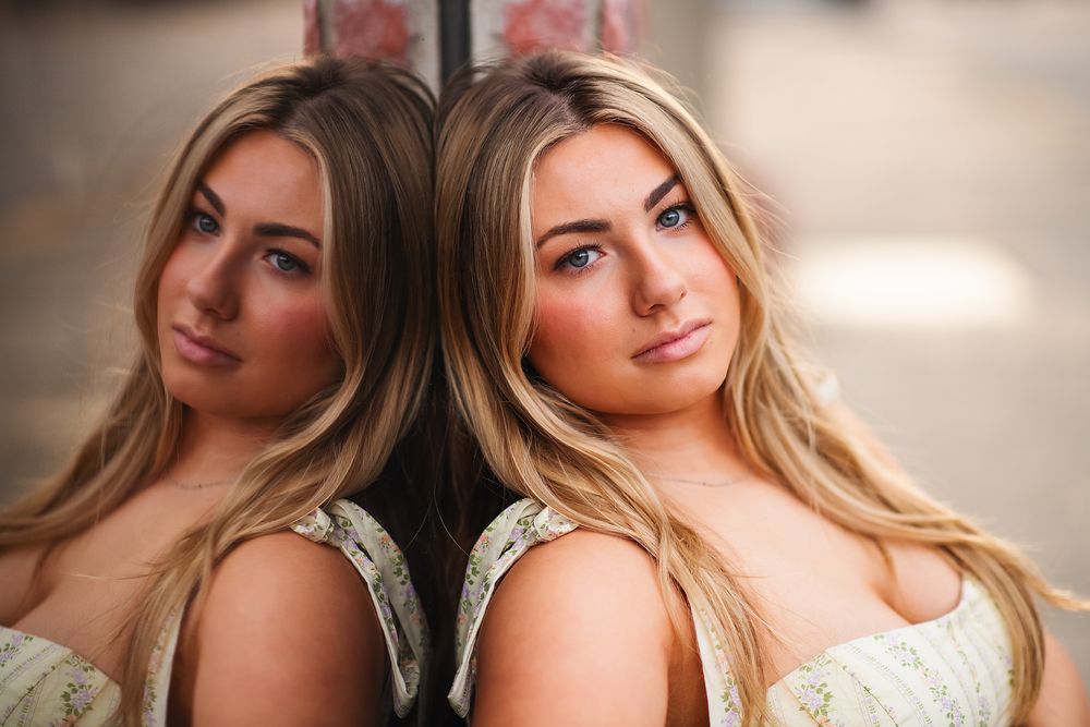 Blonde woman posing with her mirror reflection against a glass surface outdoors.