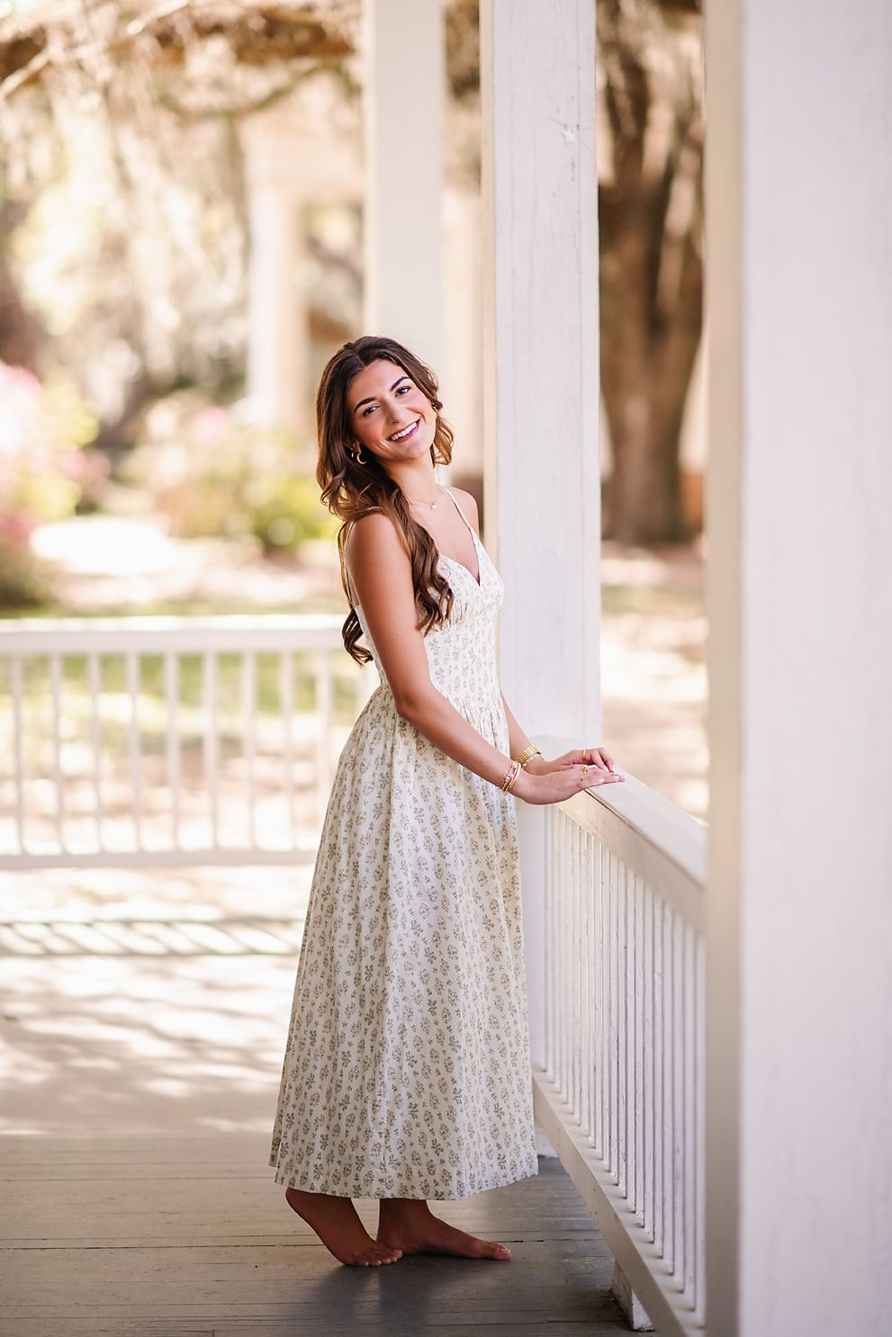 Smiling young woman in floral maxi dress leaning on white porch railing outdoors.