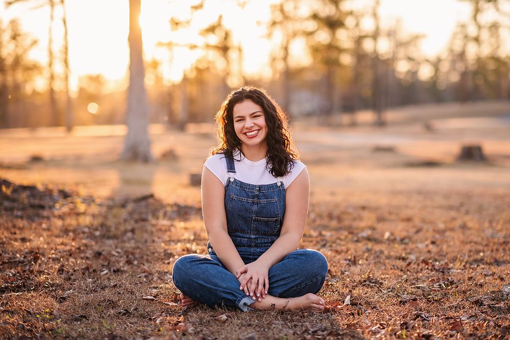 Smiling young woman in denim overalls sitting cross-legged on ground at golden hour.