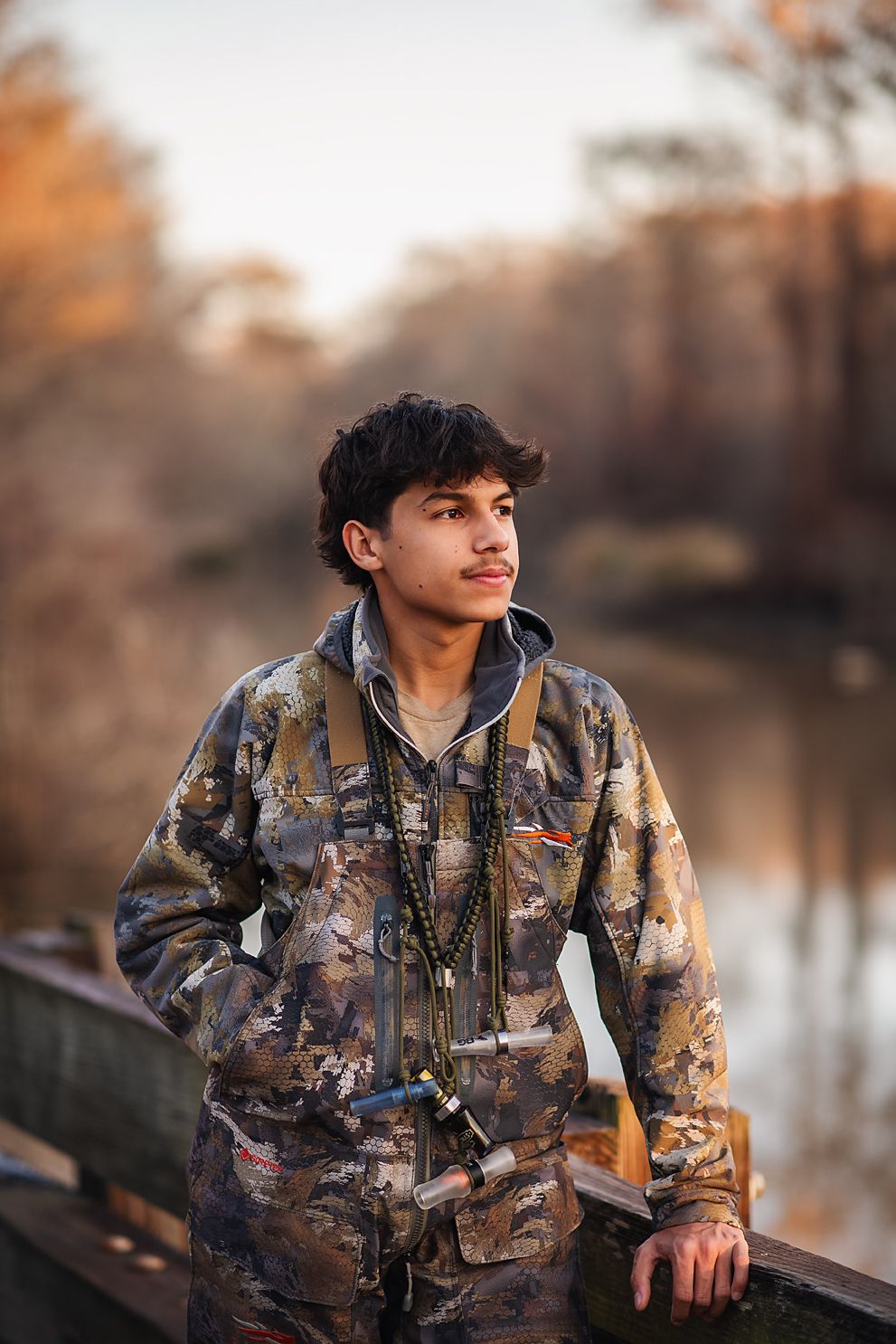 Teen boy in camo gear leaning on bridge railing near autumn lake.