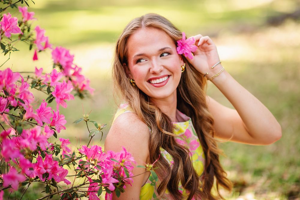 Smiling young woman holding pink flower near ear surrounded by blooming azalea bushes.